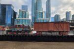 A diesel locomotive hauls a brown wooden boxcar across a railway turntable bridge. Glass highrise condos can be seen in the background