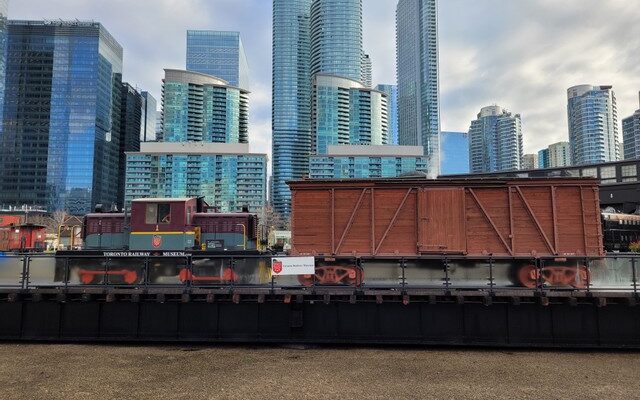 A diesel locomotive hauls a brown wooden boxcar across a railway turntable bridge. Glass highrise condos can be seen in the background