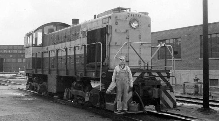 A locomotive engineer wearing coveralls poses on the front of a diesel locomotive.